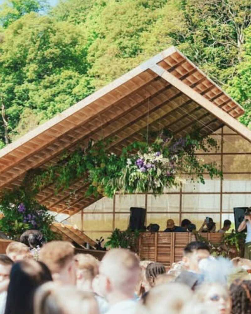 A hanging floral installation under a timber pavilion roof, with woodland behind
